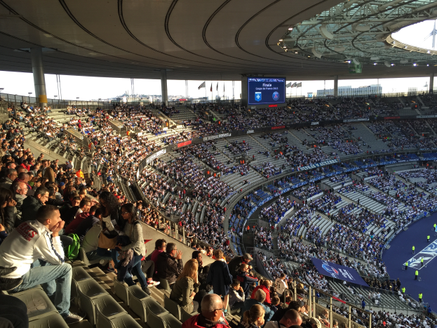 Eine große Menschenmenge sitzt im Allianz Stadion in München, Deutschland, bei einem Fussballspiel, mit einer Bühne auf der rechten Seite, Fahnen, Mästängen und einem Bildschirm im Hintergrund und dem Himmel oben sichtbar.
