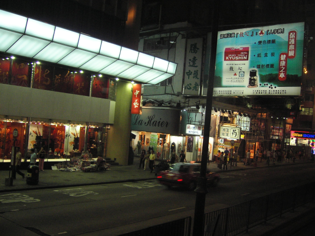 Eine nächtliche Stadtstraße mit einem Auto, das an beleuchteten Gebäuden, Geschäften und Fußgängern auf dem Bürgersteig vorbeifährt, unter einem dunklen Himmel.