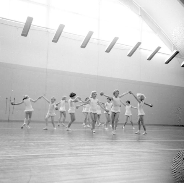 Schwarzes Bild von jungen Mädchen, die Hand in Hand tanzen und einen Ball auf der rechten Seite, Deckenlichter und ein wandmontiertes Objekt im Hintergrund.