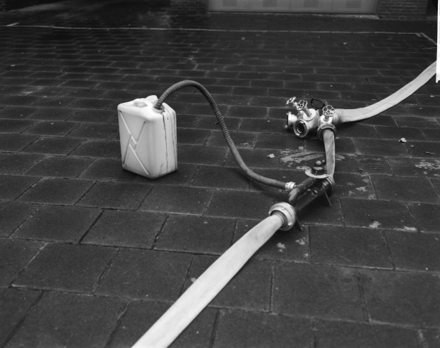 Schwarz-weißes Foto eines Gasbehälters mit einem Schlauch, der vor einem Gebäude mit einer Tür im Hintergrund auf dem Boden steht.