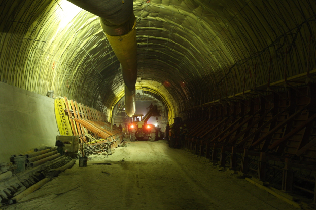 Baustelle mit einem großen Tunnel, Fahrzeugen, verstreuten hölzernen Gegenständen, Rohren, einer Wand auf der linken Seite, Überkopfrohren und beleuchteten Tunnel-Lampen im Hintergrund.