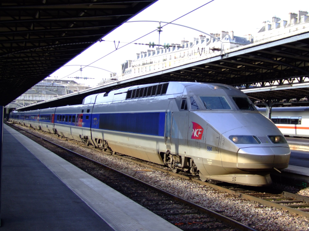 Hohe-Geschwindigkeitszug fährt auf Schienen neben einem Bahnsteig mit Gebäuden und einem klaren blauen Himmel im Hintergrund.