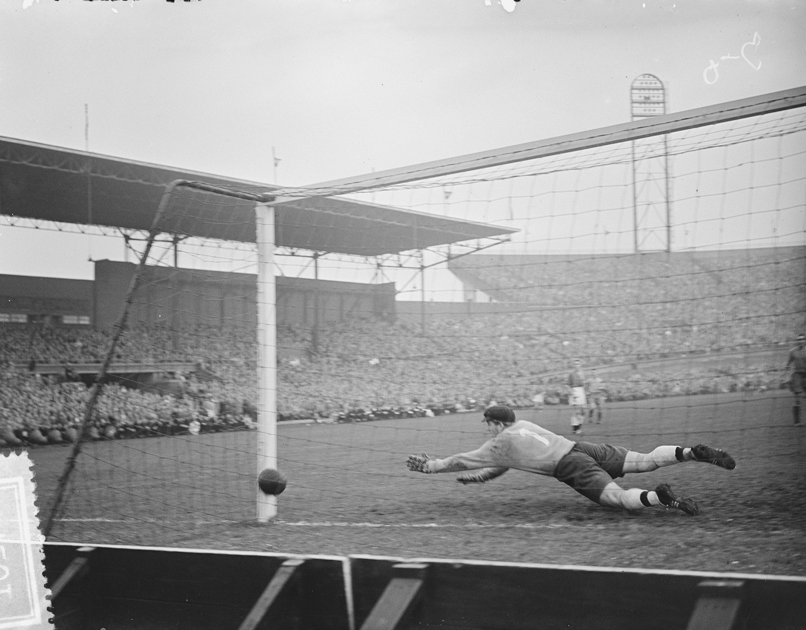 Schwarzes und weißes Foto eines Fußballspielers in Uniform, der einen Ball vor einem vollen Stadion fängt.