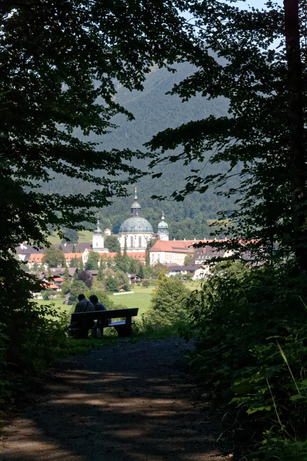 Zwei Personen sitzen auf einer Bank in einem Wald, mit Gebäuden, Bäumen und einem klaren blauen Himmel im Hintergrund.