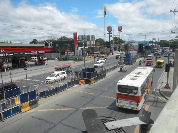 Vollgestopfte Stadtstraße mit starkem Verkehr neben einer Tankstelle, mit Fahrzeugen, Geländern, Laternen, Strommasten mit Kabeln, Schildern, Gebäuden, Bäumen und einem bewölkten Himmel.