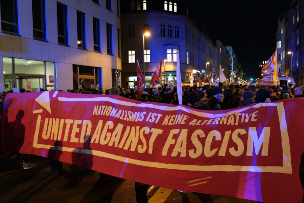 Eine Gruppe von Menschen marschiert auf einer Straße, die ein Banner mit der Aufschrift 'Einheit gegen Faschismus' trägt, mit Gebäuden, Laternen und Schildern an der Straße unter einem sichtbaren Himmel.