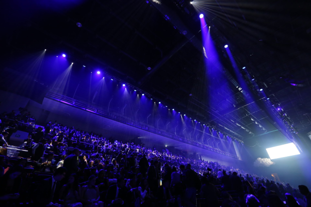 Große Menschenmenge in einem abgedunkelten Konzertsaal vor einer Bühne mit Deckenleuchten und einem Bildschirm auf der rechten Seite.