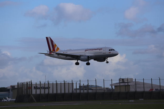 Germanwings Airbus A320-200 beim Start vom Frankfurt Airport, mit Gras darunter, einem Zaun im Vordergrund, Gebäuden und Bäumen im Hintergrund und einer bewölkten Himmel über dem Flugzeug.