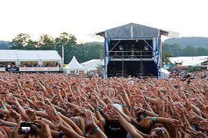 Eine große Menschenmenge auf einem Musikfestival mit erhobenen Händen und Mobiltelefonen, eine Bühne im Hintergrund, weiße Zelte, Bäume und einen klaren blauen Himmel.