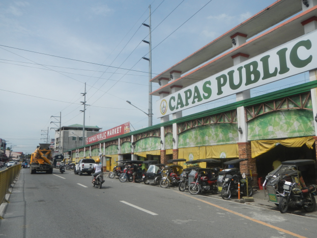 Eine belebte Stadtstraße mit Fahrzeugen, einem Geländer auf der linken Seite, Gebäuden, Pfählen, Lampen, Schildern und einigen Menschen bei einem bewölkten Himmel, mit einem Gebäude mit der Aufschrift "Capas Public".