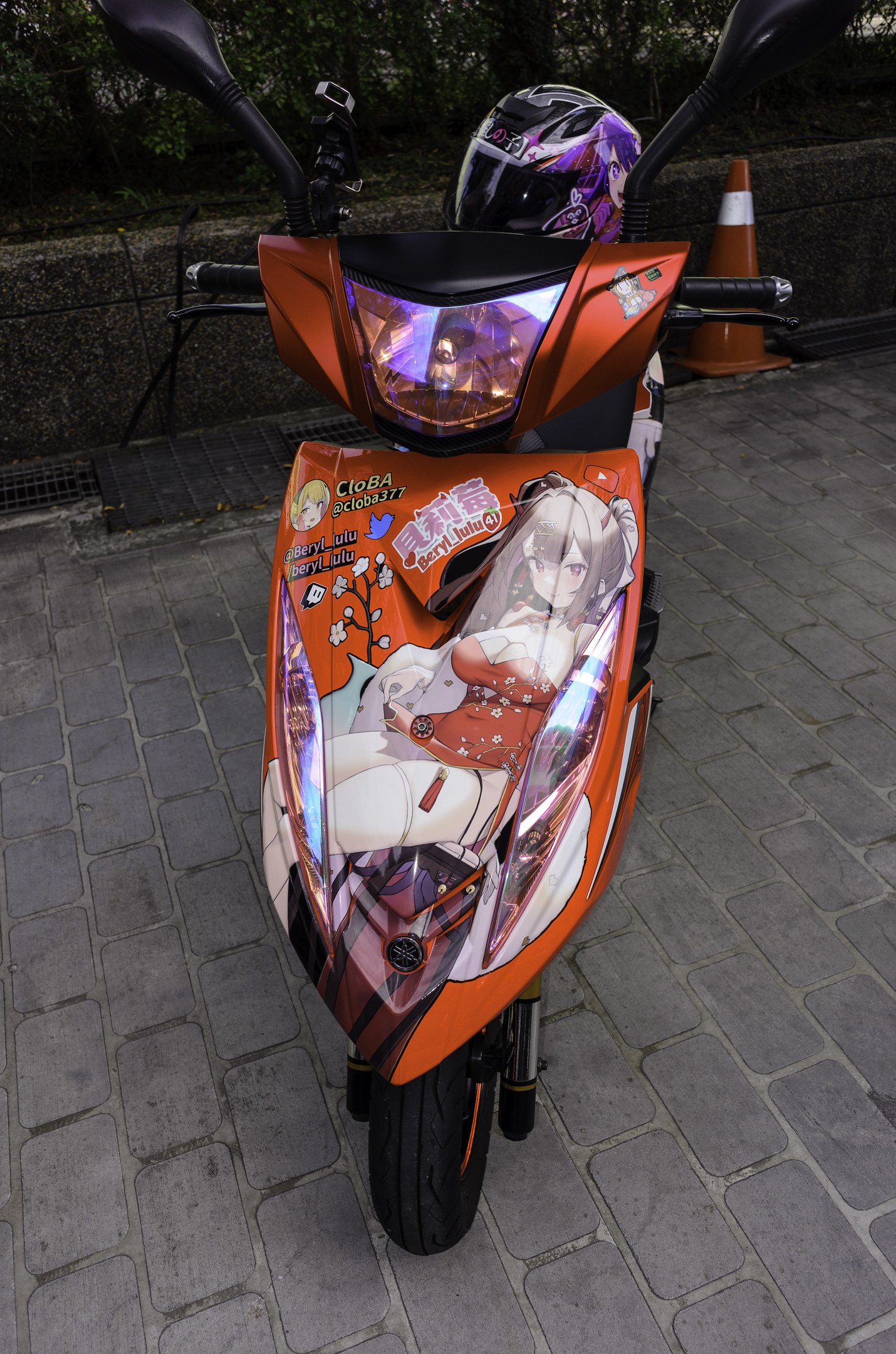 Ein oranges E-Scooter auf einer Straße mit einem Helm darauf, einer Verkehrskegel, einer Wand und Bäumen im Hintergrund.