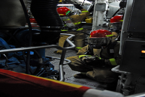 Feuerwehrleute in Helmen und Schutzausrüstung arbeiten an einem Löschfahrzeug mit Rohren und anderen Gegenständen im Hintergrund, unter einem Licht an der Spitze.