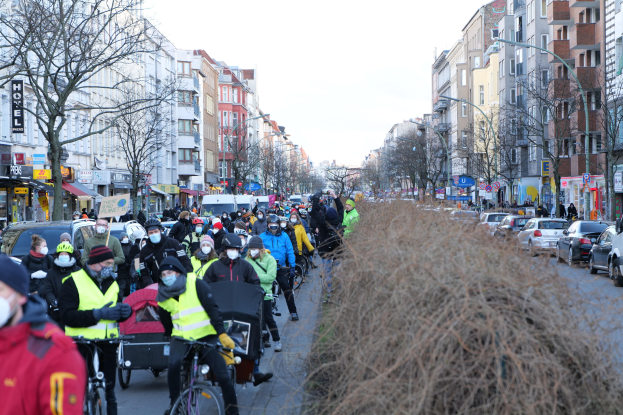 Eine große Gruppe von Menschen in Masken und Sicherheitswesten auf Fahrrädern eine straßenläuflich mit Bäumen, Gebäuden, Laternen, Texttafeln, Fahrzeugen, trockenem Gras und einem klaren blauen Himmel.