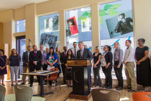 Eine Gruppe von Menschen steht vor einem Rednerpult mit Mikrofon, umgeben von Tischen, Stühlen und Papieren, in einem Raum mit Glasfenstern, die Plakate zeigen, und einer beleuchteten Decke, was auf eine Schuleröffnung hindeutet.