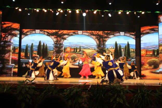 Eine Gruppe von Menschen in bunten Kostümen performs auf einer Bühne umgeben von Pflanzen unten und einem Bildschirm im Hintergrund, beleuchtet von Lampen oben.