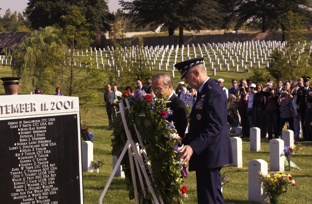 Eine Gruppe von Menschen steht um ein Grab auf einem Friedhof, wobei ein Mann in einer Militäruniform einen Kranz darauflegt, umgeben von Blumenbouquets, einer Tafel mit Text und Bäumen unter einem bewölkten Himmel.