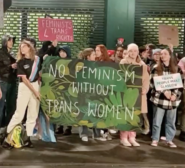 Eine Gruppe von Menschen hält ein Transparent mit der Aufschrift "Kein Feminismus ohne Trans-Frauen" und Schilder mit verschiedenen Botschaften in der Hand und steht an einem öffentlichen Ort mit einer Wand und einem Rollladen im Hintergrund.