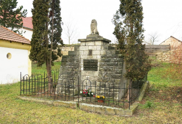 Ein steinerner Gedenkstein auf einer Rasenfläche, umgeben von einem Metallzaun und geschmückt mit Blumensträußen, mit Bäumen, Häusern und einer Wand im Hintergrund unter einem klaren blauen Himmel, als Holocaust-Gedenkstätte in Sibiu, Rumänien.