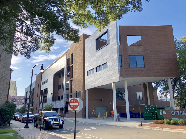 Großes Rathausgebäude mit vielen Fenstern, umgeben von Straßeninfrastruktur, Fahrzeugen, Grünflächen und einem bewölkten Himmel.