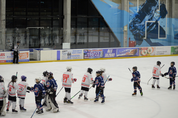 Junge Menschen in Helmen und Hockey-Ausrüstung spielen Eis-hockey auf einer Indoor-Eisfläche, mit Bannern, Säulen und Glaswänden im Hintergrund.