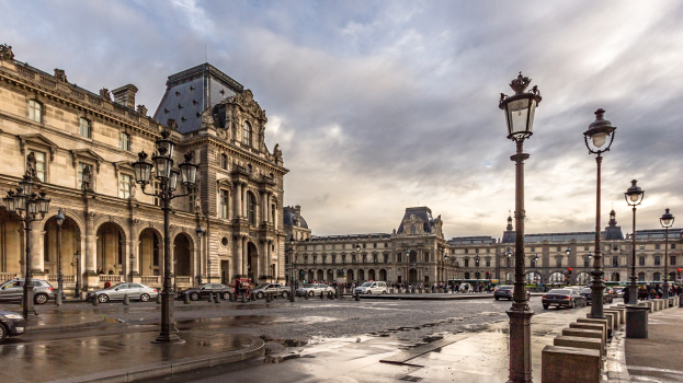 Außenansicht des Louvre-Museums in Paris, Frankreich, mit seiner ikonischen Architektur, Laternenpfählen, Fahrzeugen, Passanten auf dem Gehweg und einer bewölkten Himmel.