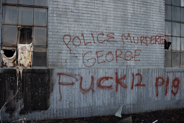 Graffiti auf einem Gebäudewand mit der Aufschrift "Polizei ermordet George Fuck 12 P19" mit Fenstern und verstreuten Gegenständen davor.